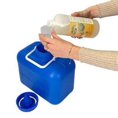 Eine Person gießt Urinbehälterzusatz für Trenntoiletten "Liquid Fresh" in einen Messbecher über einem großen blauen Kunststoffbehälter mit weißem Griff und offenem Deckel.